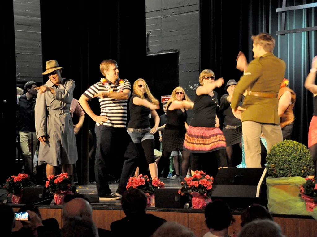 Tanzgruppe in Mänteln und Hüten auf der Bühne.